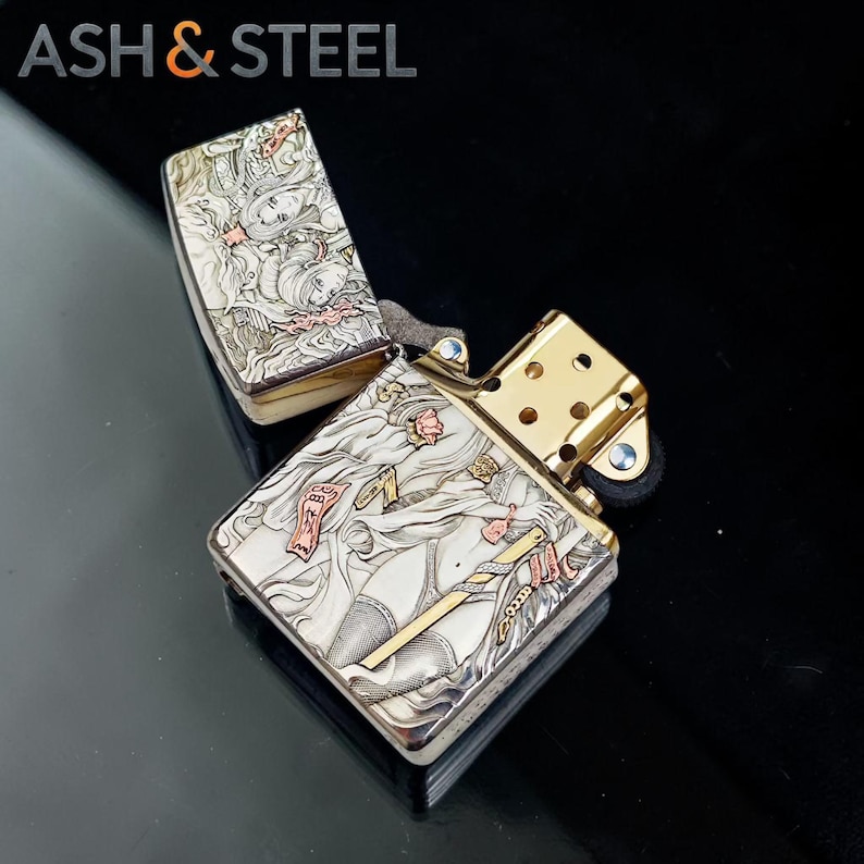 May include: A silver and gold lighter with an open lid, featuring an engraved design of a woman in a flowing gown. The lighter has a gold-colored top and a black base. The text "ASH & STEEL" is visible in the top left corner.