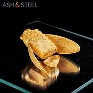 Puede incluir: Un encendedor dorado con forma de cigarra, con detalles intrincados en el cuerpo y las alas. El encendedor es de metal y tiene una tapa con bisagras. La marca "ASH & STEEL" es visible en la esquina superior izquierda.