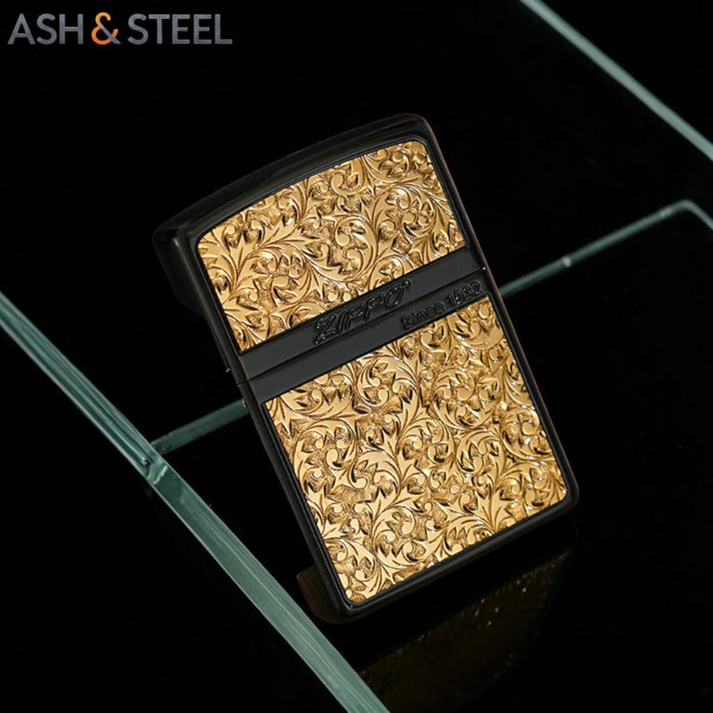 May include: Black and gold Zippo lighter with an ornate floral pattern. The lighter has a black band across the middle with the Zippo logo and "Since 1932" printed on it. The lighter is set against a black background.