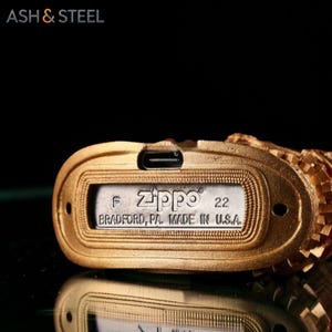 May include: Close-up of a gold-colored Zippo lighter. The lighter's base features the Zippo logo, the number 22, and the text "BRADFORD, PA. MADE IN U.S.A." The brand name "ASH & STEEL" is visible in the top left corner.