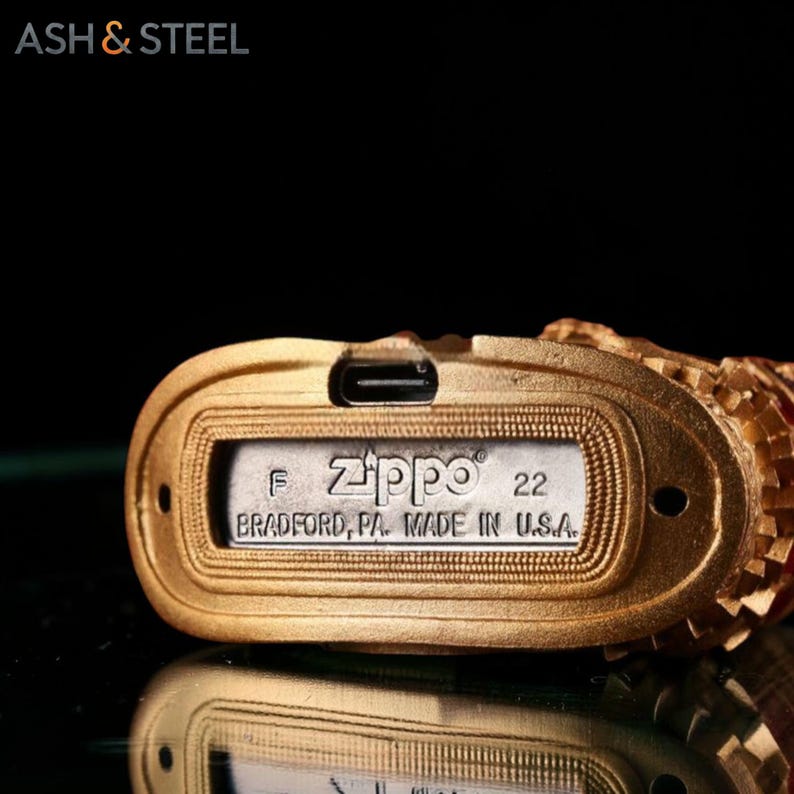 May include: Close-up of a gold-colored Zippo lighter. The lighter's base features the Zippo logo, the number 22, and the text "BRADFORD, PA. MADE IN U.S.A." The brand name "ASH & STEEL" is visible in the top left corner.