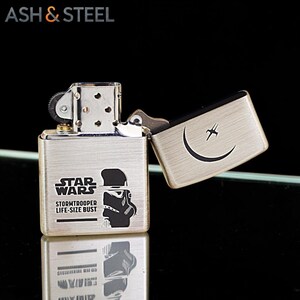 K&ouml;nnte beinhalten: Ein silbernes Metallfeuerzeug mit dem Star Wars-Logo und einer Stormtrooper-Grafik. Das Feuerzeug ist ge&ouml;ffnet und zeigt ein Design aus Halbmond und Stern. Der Text "ASH & STEEL" befindet sich oben.