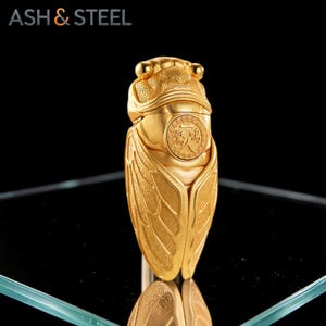Puede incluir: Figura de cigarra dorada con detalles intrincados, incluyendo alas grabadas y un emblema circular. La figura se exhibe sobre una superficie reflectante, con el texto "ASH & STEEL" en la esquina superior izquierda.