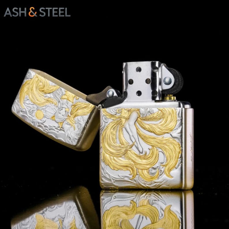 May include: A silver and gold lighter with an engraved design. The lighter features a detailed relief of a woman with flowing hair. The text "ASH & STEEL" is in the upper left corner. The lighter is open, revealing the interior.