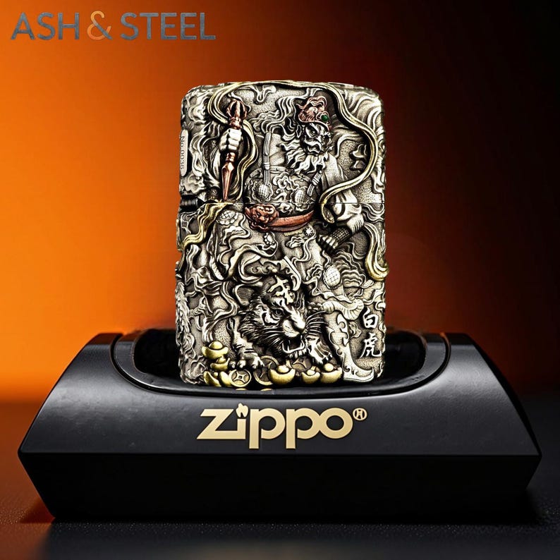 May include: A detailed, silver-toned Zippo lighter with an intricate relief design featuring a tiger and other figures. The lighter is displayed on a black stand with the Zippo logo in gold. The text "ASH & STEEL" is visible at the top.
