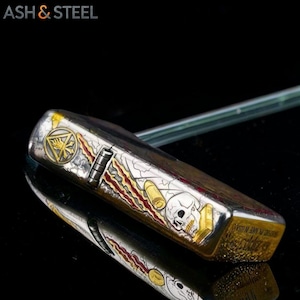 May include: A silver and gold metal lighter with a skull and bullet design. The lighter has a detailed graphic with a skull, bullets, and a gold emblem. The text "ASH & STEEL" is in the upper left corner.