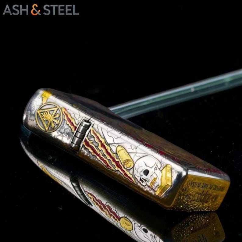 May include: A silver and gold metal lighter with a skull and bullet design. The lighter has a detailed graphic with a skull, bullets, and a gold emblem. The text "ASH & STEEL" is in the upper left corner.