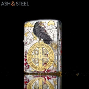 May include: Silver lighter with a detailed raven design. The raven is perched on a gold-colored emblem with the text "CSMDB". The lighter has a decorative spiderweb and a sun and cloud design.