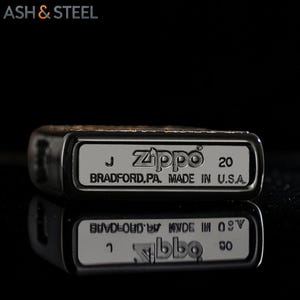 May include: Close-up of a Zippo lighter with the text "Zippo" and "BRADFORD.PA. MADE IN U.S.A." The lighter is silver with a black border. The lighter's reflection is visible below it.