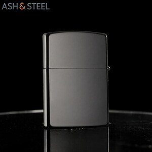 May include: A shiny, dark gray lighter with a rectangular shape. The lighter has a hinged lid and a smooth, reflective surface. The text "ASH & STEEL" is in the upper left corner.