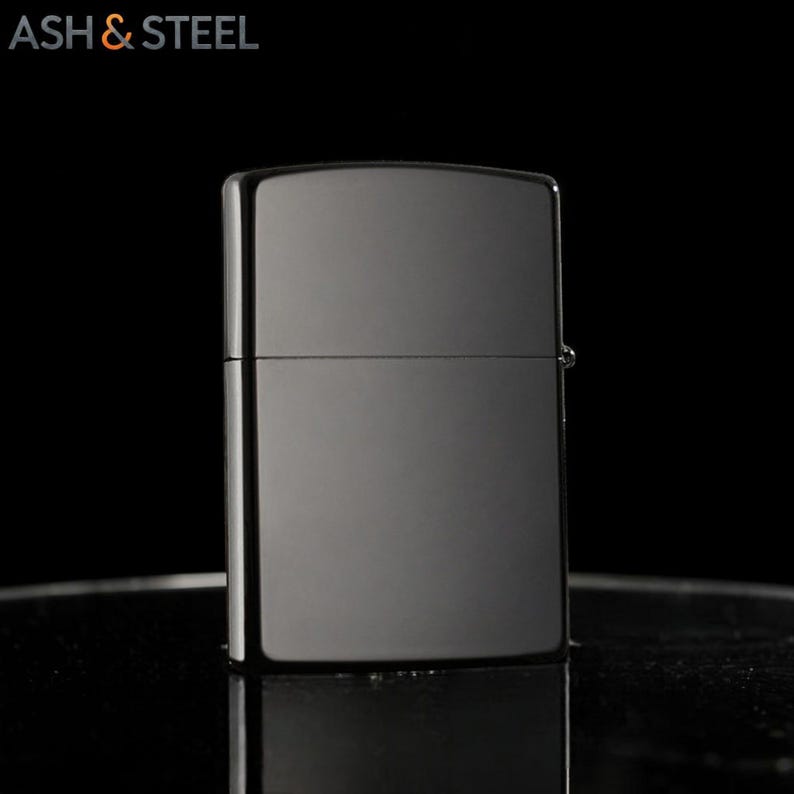 May include: A shiny, dark gray lighter with a rectangular shape. The lighter has a hinged lid and a smooth, reflective surface. The text "ASH & STEEL" is in the upper left corner.