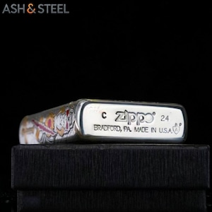 May include: A silver Zippo lighter with a colorful design on the side. The lighter has the text "Zippo 24" and "BRADFORD, PA. MADE IN U.S.A." printed on it. The lighter is resting on a black box.