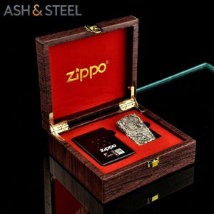 May include: A Zippo lighter and a black card are displayed inside a wooden box with a red velvet lining. The lighter has an intricate design. The box is branded with the text "ASH & STEEL" and "Zippo".