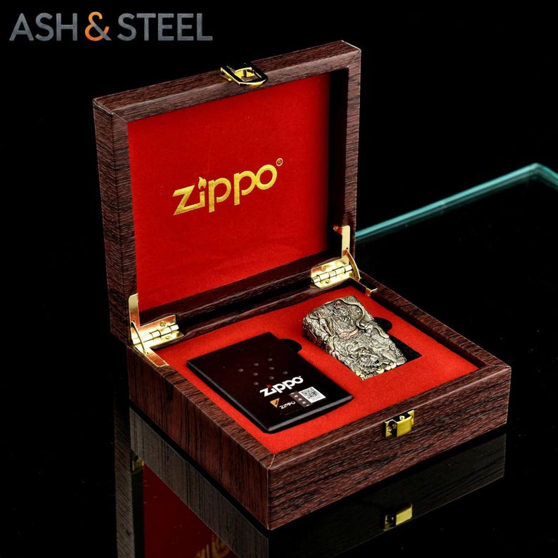 May include: A Zippo lighter and a black card are displayed inside a wooden box with a red velvet lining. The lighter has an intricate design. The box is branded with the text "ASH & STEEL" and "Zippo".