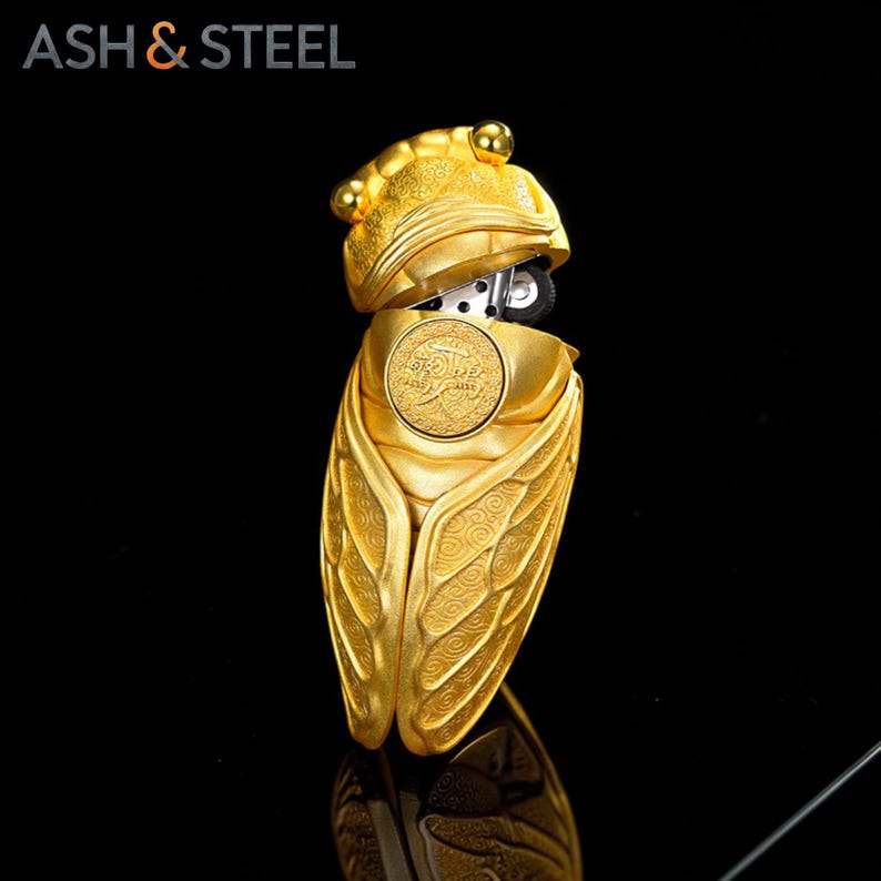 Puede incluir: Un encendedor dorado con forma de cigarra. El encendedor est&aacute; abierto, revelando el mecanismo de encendido. Las alas y el cuerpo de la cigarra est&aacute;n detallados con patrones en relieve. El texto "ASH & STEEL" est&aacute; en la esquina superior izquierda.