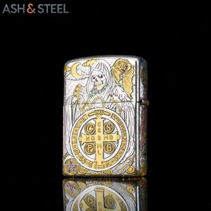 May include: Silver and gold-toned metal lighter with a detailed design. The lighter features a skull with a hooded cloak, a cross, and the text "ASH & STEEL". The design includes intricate engravings and a decorative pattern.
