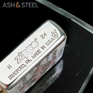 May include: Close-up of a silver Zippo lighter. The lighter is marked with the Zippo logo, the number 24, and "MADE IN U.S.A." The text "BRADFORD, PA" is also visible. The lighter is on a reflective surface.