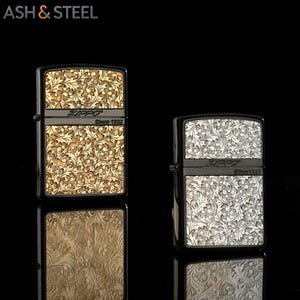 May include: Two Zippo lighters with ornate floral patterns. One lighter has a gold finish, the other a silver finish. Both lighters have black frames and the Zippo logo with "Since 1932" printed on them. The lighters are displayed on a reflective black surface.