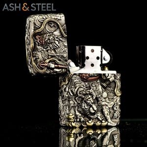May include: A silver-colored metal lighter with intricate relief carvings of a tiger and dragon, with gold and red accents. The lighter is open, revealing the internal mechanism. The text "ASH & STEEL" is visible at the top.