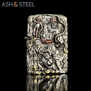 May include: A silver-colored lighter with intricate relief carvings. The design features a detailed depiction of a warrior figure and a tiger, with gold and copper accents. The text "ASH & STEEL" is at the top.