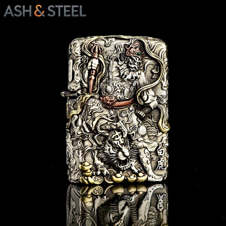 May include: A silver-colored lighter with intricate relief carvings. The design features a detailed depiction of a warrior figure and a tiger, with gold and copper accents. The text "ASH & STEEL" is at the top.
