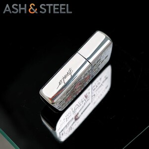 May include: A silver-colored lighter with the word "Breaker" in cursive script. The lighter has a rectangular shape with a shiny, reflective surface. The top of the lighter is a solid silver color. The brand name "ASH & STEEL" is in the top left corner.