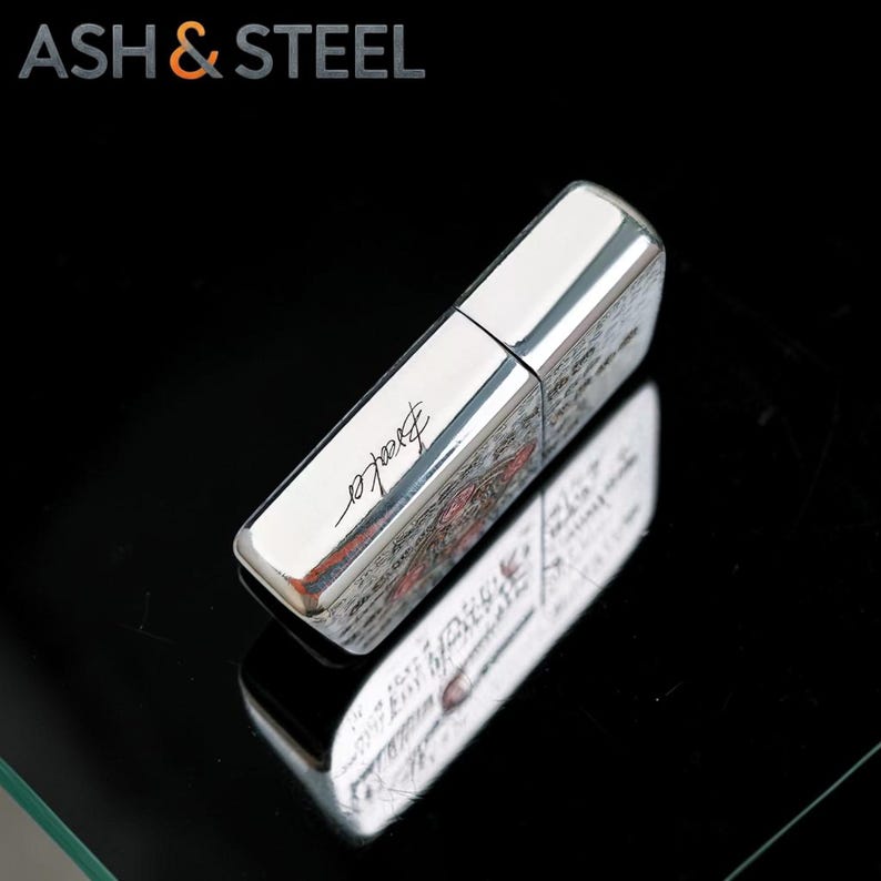 May include: A silver-colored lighter with the word "Breaker" in cursive script. The lighter has a rectangular shape with a shiny, reflective surface. The top of the lighter is a solid silver color. The brand name "ASH & STEEL" is in the top left corner.