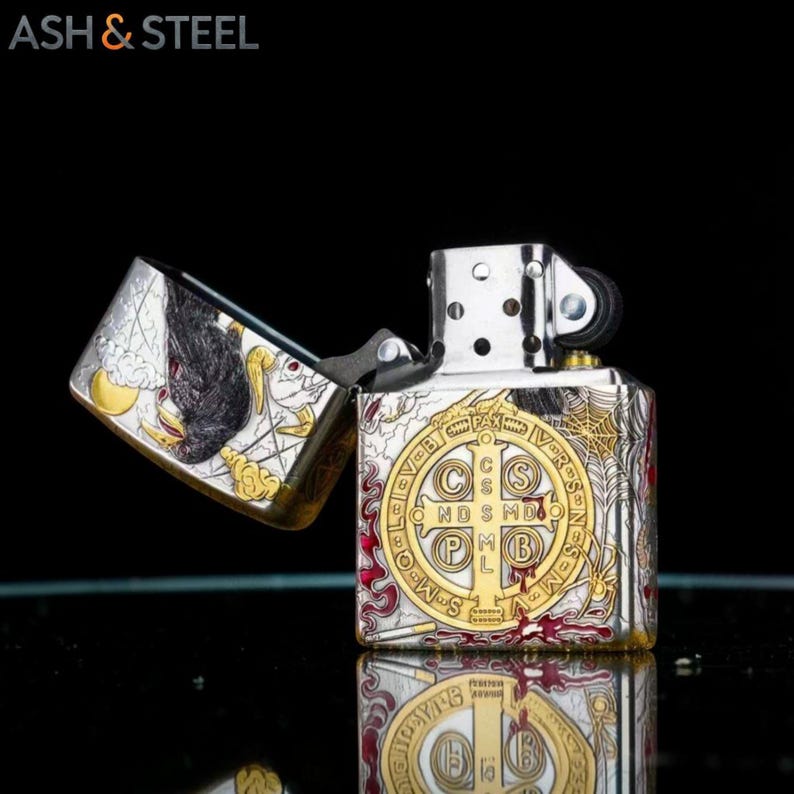 May include: Silver and gold-toned metal lighter with an open lid. The lighter features an intricate design with religious symbols and a raven illustration. The text "ASH & STEEL" is in the upper left corner.
