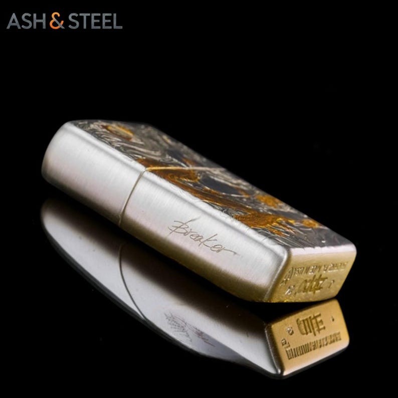 May include: A silver and gold lighter with the word "Breaker" in script. The top of the lighter has a swirling gold and black design. The bottom of the lighter is gold with engraved text. The brand name "ASH & STEEL" is in the top left corner.
