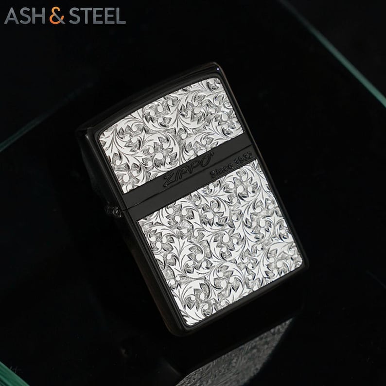 May include: Black Zippo lighter with a silver floral pattern. The lighter has a black band across the middle with the Zippo logo and "Since 1932" printed on it. The brand name "ASH & STEEL" is in the top left corner.