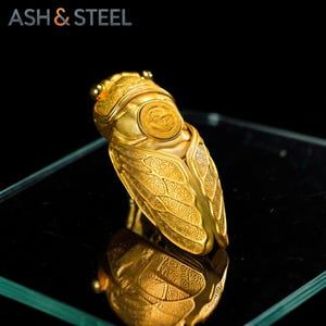 Puede incluir: Un objeto dorado con forma de cigarra con intrincados detalles en las alas. La pieza inspirada en insectos tiene una superficie reflectante y est&aacute; sobre un fondo negro. El texto "ASH & STEEL" es visible en la esquina superior izquierda.