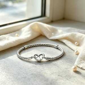 May include: A silver-toned bracelet featuring a woven, chain-link texture and a central heart-shaped clasp. The bracelet is constructed with a flexible, braided metal design that connects to a smooth, polished heart-shaped closure mechanism.