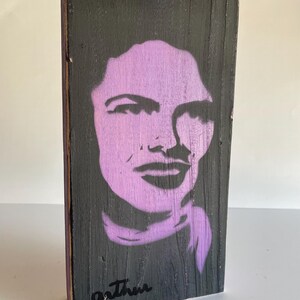 Peut inclure: Une &oelig;uvre d'art en bois avec un portrait au pochoir violet sur fond noir. Le portrait repr&eacute;sente le visage d'une femme. La signature de l'artiste, "Arthur", est en noir en bas. Le bois a un aspect rustique et textur&eacute;.