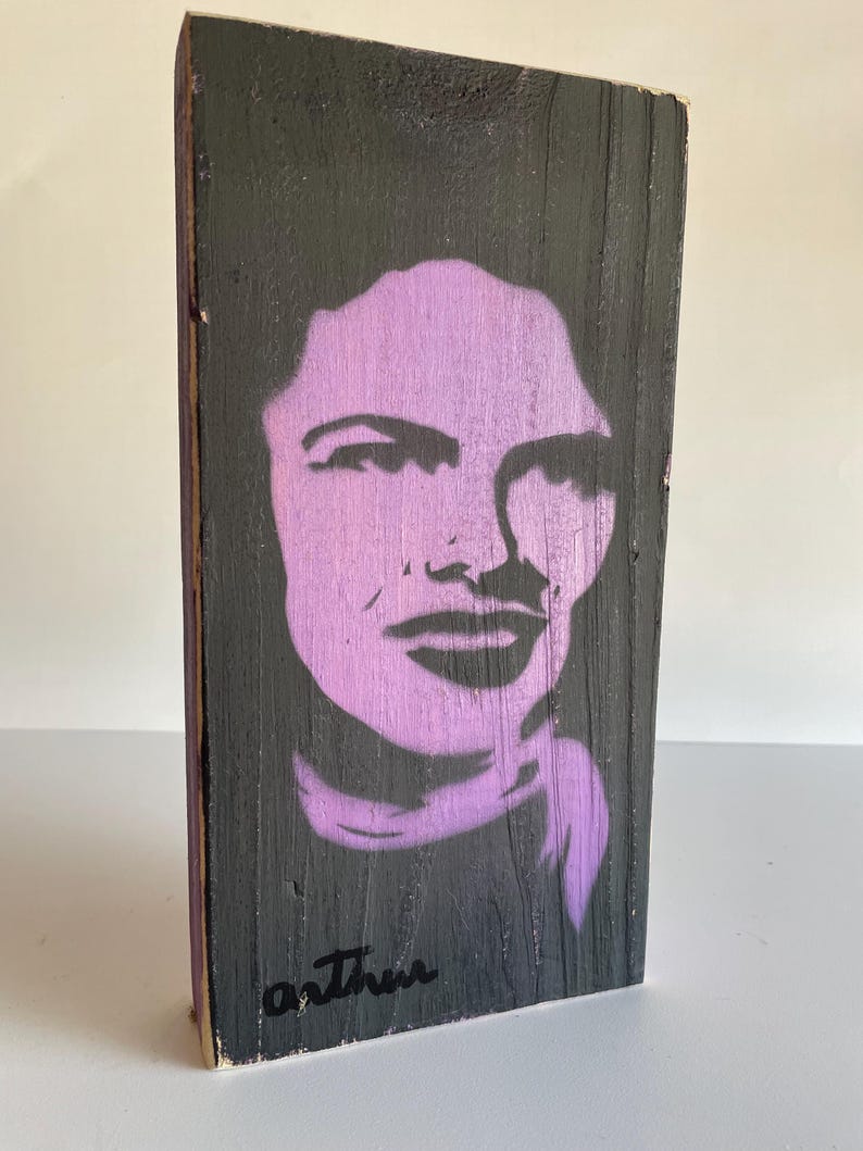 Peut inclure: Une &oelig;uvre d'art en bois avec un portrait au pochoir violet sur fond noir. Le portrait repr&eacute;sente le visage d'une femme. La signature de l'artiste, "Arthur", est en noir en bas. Le bois a un aspect rustique et textur&eacute;.