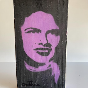May include: A rectangular wooden block with a black painted surface. A purple stencil portrait of a person's face is centered on the block. The signature "o. Thru" is in black ink at the bottom.