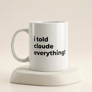 May include: White ceramic mug with a curved handle, printed with the text "i told claude everything!" in black. The mug sits on a light-coloured, triangular stand. The background is a plain, neutral colour.