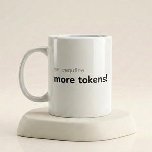 Puede incluir: Taza de cerámica blanca con asa curva, con el texto en negro "we require more tokens!". La taza está sobre una plataforma beige claro, de forma triangular. El fondo es de un tono suave y neutro.