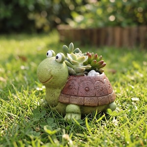 Könnte beinhalten: Ein schildkrötenförmiger Blumentopf, bemalt in Grün- und Brauntönen, steht im Gras. Der Panzer der Schildkröte beherbergt eine Auswahl an Sukkulenten. Der Topf hat ein lächelndes Gesicht und ist ein bezaubernder Gartenschmuck. Etwa 20 cm lang.