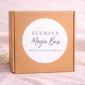 Könnte beinhalten: Eine quadratische, hellbraune Pappschachtel mit einem weißen, kreisförmigen Etikett. Auf dem Etikett steht "ELUNIVA Magic Box" in einer Schriftart, mit zusätzlichem Text darunter. Die Box ist geschlossen und liegt auf einer hellrosa Oberfläche.