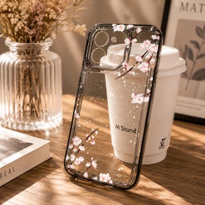 May include: A clear smartphone case featuring a delicate cherry blossom branch design with pink flowers and subtle white glitter accents. The case has a dark border and precise cutouts for the camera lenses, providing a transparent protective cover.