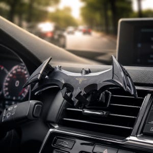 May include: A black car air vent clip shaped like a stylized bat emblem with sharp, angular wings and a small gold-colored triangular logo centered on the front. The accessory is securely attached to the horizontal slats of a vehicle dashboard vent.