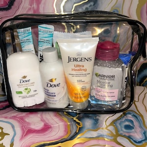 Könnte beinhalten: Eine transparente Reisetasche mit verschiedenen Toilettenartikeln. Enthalten sind Dove Deodorant, Dove Bodylotion, Jergens Ultra Healing Feuchtigkeitscreme (59 ml), Garnier Mizellen Reinigungswasser und ChapStick. Der Hintergrund ist ein farbenfrohes, abstraktes Muster.