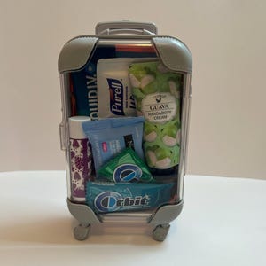 K&ouml;nnte beinhalten: Ein durchsichtiger Miniaturkoffer gef&uuml;llt mit Toilettenartikeln in Reisegr&ouml;&szlig;e. Zu den Artikeln geh&ouml;ren H&auml;ndedesinfektionsmittel, Handcreme, Lippenbalsam und Kaugummi. Auf der Handcreme-Verpackung steht "GUAVA HAND&BODY CREAM". Auf der Kaugummi-Verpackung steht "Orbit WINTERMINT GUM".