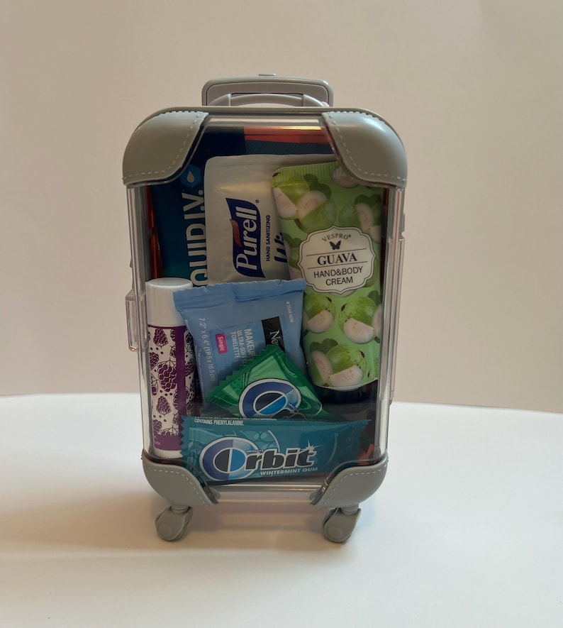 K&ouml;nnte beinhalten: Ein durchsichtiger Miniaturkoffer gef&uuml;llt mit Toilettenartikeln in Reisegr&ouml;&szlig;e. Zu den Artikeln geh&ouml;ren H&auml;ndedesinfektionsmittel, Handcreme, Lippenbalsam und Kaugummi. Auf der Handcreme-Verpackung steht "GUAVA HAND&BODY CREAM". Auf der Kaugummi-Verpackung steht "Orbit WINTERMINT GUM".