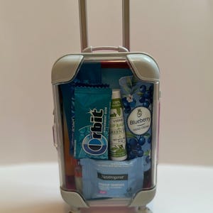 K&ouml;nnte beinhalten: Ein kleiner, durchsichtiger Koffer mit ausziehbarem Griff und R&auml;dern, gef&uuml;llt mit Toilettenartikeln in Reisegr&ouml;&szlig;e. Enth&auml;lt Orbit Wintermint-Kaugummi, Neutrogena Make-up-Entferner-T&uuml;cher, Visrie Lippenbalsam und Blaubeer-Handcreme.