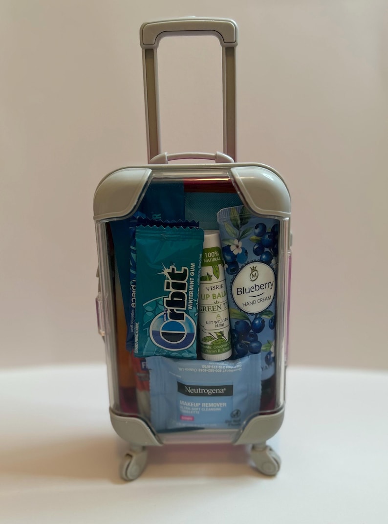 K&ouml;nnte beinhalten: Ein kleiner, durchsichtiger Koffer mit ausziehbarem Griff und R&auml;dern, gef&uuml;llt mit Toilettenartikeln in Reisegr&ouml;&szlig;e. Enth&auml;lt Orbit Wintermint-Kaugummi, Neutrogena Make-up-Entferner-T&uuml;cher, Visrie Lippenbalsam und Blaubeer-Handcreme.