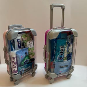 K&ouml;nnte beinhalten: Zwei Miniaturkoffer, einer rosa und einer magenta, gef&uuml;llt mit Toilettenartikeln und Snacks in Reisegr&ouml;&szlig;e. Die Koffer haben graue Akzente, transparente Seiten und ausziehbare Griffe. Enth&auml;lt Kaugummi, Handcreme und Lippenbalsam.
