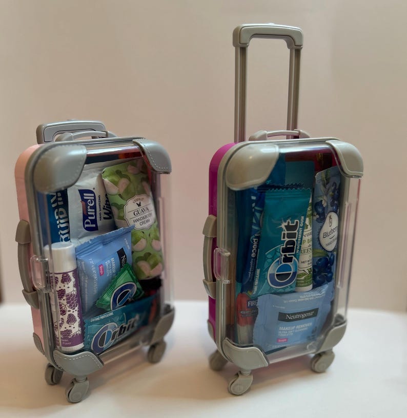 K&ouml;nnte beinhalten: Zwei Miniaturkoffer, einer rosa und einer magenta, gef&uuml;llt mit Toilettenartikeln und Snacks in Reisegr&ouml;&szlig;e. Die Koffer haben graue Akzente, transparente Seiten und ausziehbare Griffe. Enth&auml;lt Kaugummi, Handcreme und Lippenbalsam.