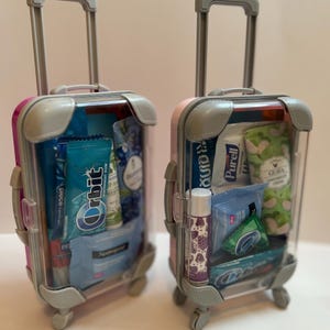 K&ouml;nnte beinhalten: Zwei transparente Miniaturkoffer mit rosa und grauen Akzenten, gef&uuml;llt mit Toilettenartikeln und Snacks in Reisegr&ouml;&szlig;e. Einer ist rosa und der andere lila. Enth&auml;lt Kaugummi, Lippenbalsam und Handcreme.