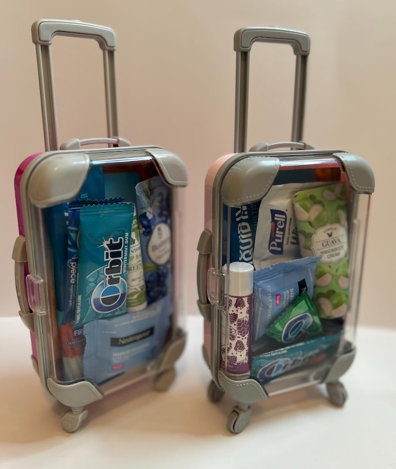 K&ouml;nnte beinhalten: Zwei transparente Miniaturkoffer mit rosa und grauen Akzenten, gef&uuml;llt mit Toilettenartikeln und Snacks in Reisegr&ouml;&szlig;e. Einer ist rosa und der andere lila. Enth&auml;lt Kaugummi, Lippenbalsam und Handcreme.